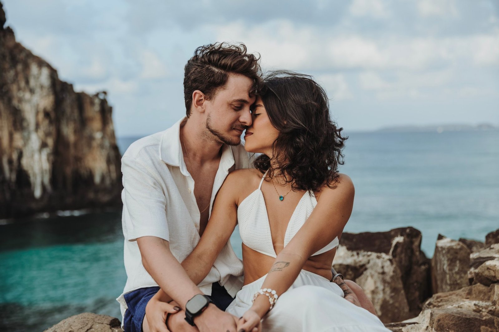 Juliana e Patrick sentados nas pedras, rosto com rosto, diante do mar de Fernando de Noronha